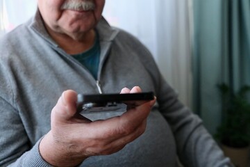 An elderly man is sitting by the window with a smartphone in his hands and talking. The smartphone is in the hands of an elderly retired man.
