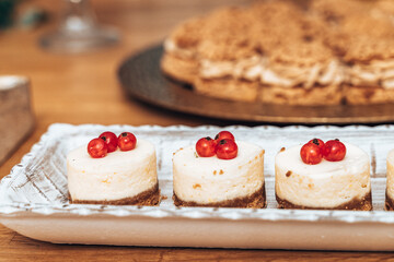 Mini cheesecakes with a smooth texture, each topped with a trio of vibrant red currants. Presented on a rustic ceramic tray, these bite-sized treats have a light, creamy finish over a crumbly crust.

