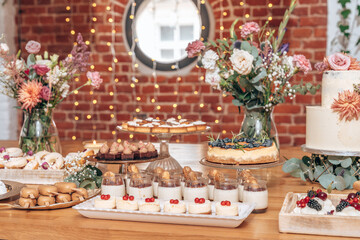 A charming dessert table display with various sweets, including mini cheesecakes, panna cotta, chocolate squares, and donuts, all set against a rustic brick wall with twinkling lights and floral arran
