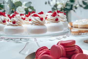 Delicate meringue desserts topped with whipped cream, fresh raspberries, and red currants, arranged elegantly on a glass stand. In the foreground, bright pink macarons add a pop of color