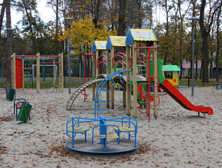 Sand covered with autumn leaves on the empty multi-colored playground with slides and small merry go round. No children at cold season at the play yard for active games 