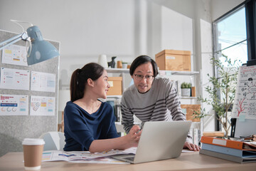 Two young Asian startup company partners discuss and brainstorm in casual small business office, marketing planning jobs on a laptop, and freelance work as E-commerce entrepreneur and coworker team.