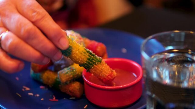 an adult's hand dipping a baked stick into jam rainbow churros, ube ice cream, strawberry caramel beautiful presentation of ice cream on a blue plate with strawberry jam in a red bowl