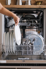 Front view from female hand removing clean plate from a loaded dishwasher. Background of modern kitchen