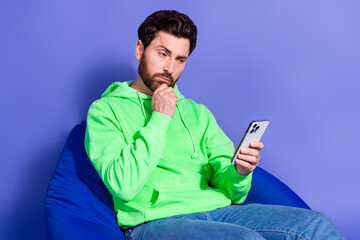 Photo portrait of pretty young male skeptical hold device sit beanbag wear trendy green outfit isolated on purple color background