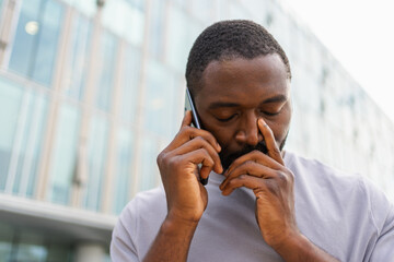 Sad tired lonely disappointed African man alone talking on smartphone on city street outdoor. Unhappy person making answering call by cellphone experiencing grief getting bad news. Stressed man