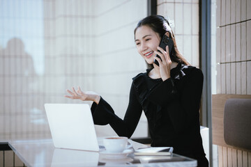 People relaxing in cafe concept. Happy face of adult Asian woman using smartphone on table with laptop computer and drink.