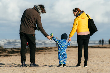 A happy family strolls along a chilly autumn beach, with their young son joyfully running ahead,...