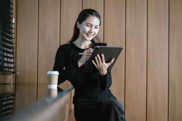 A beautiful young Asian woman in casual clothes sits at a table in a coffee shop with her digital...