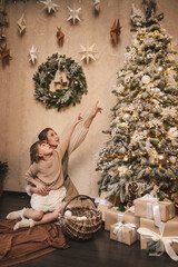 Happy mother and daughter playing near christmas decorated tree. beige and golden new year decorations, bokeh and pine wreath. Toned grain noise vintage image