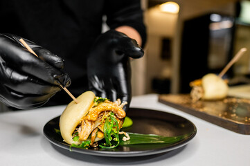 A chef’s hands in black gloves carefully plating an Asian fusion bao bun with savory fillings on...