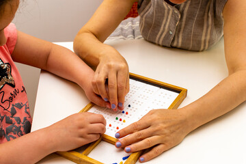 The hands of a girl and the hands of a teacher in close-up. Braille board for learning alphabet.