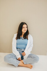 A woman in a white shirt and jeans stands by a wall, showing a stylish and understated look. The light background and natural pose give the shot a modern feel.