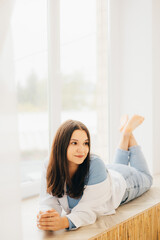 girl lying on her stomach on the windowsill, pensive look, slight smile in a white shirt and light jeans