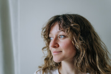 Young woman with curly brown hair and blue eyes is gazing thoughtfully into the distance, her face...