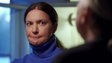 Adult 44 year old woman having lively conversation with elderly woman sitting at table in living room. Woman with anxious, tense face listens and talks.