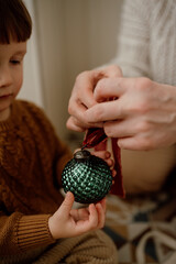 Grandmother and grandson in knitted sweaters tie a velvet ribbon on a green vintage Christmas tree ball. Preparation for Christmas
