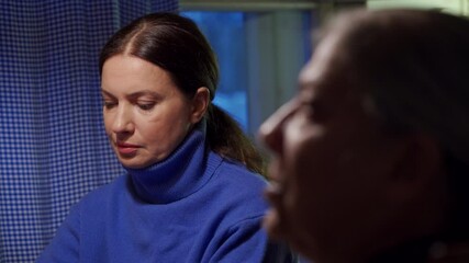 Adult 44 year old woman having lively conversation with elderly woman sitting at table in living room. Woman with tense face listens, speaks, looks at the phone screen.