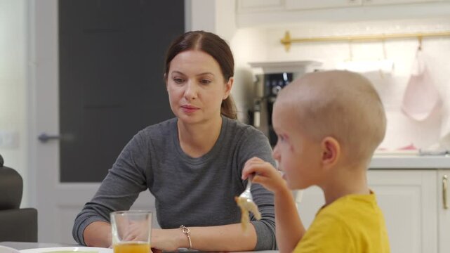 Adult 44 year old woman with tired face watches her child at the kitchen table. Woman talks to the boy, laughing and persuading her son to eat lunch.