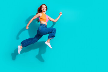 Young woman with red hair jumps energetically against a vibrant teal background wearing a summer casual outfit and flared jeans