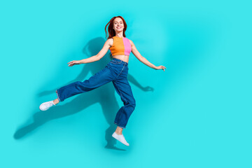 Joyful young woman with red hair leaping in the air in a summer outfit on a teal background