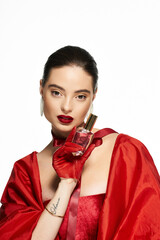 A stunning woman showcases a perfume bottle while dressed in luxurious red fabric.