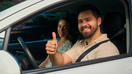 Happy Caucasian family couple sitting inside automobile man male guy driver fasten seat belt smiling showing thumb up gesture woman female girl car passenger driving safety rental service test drive