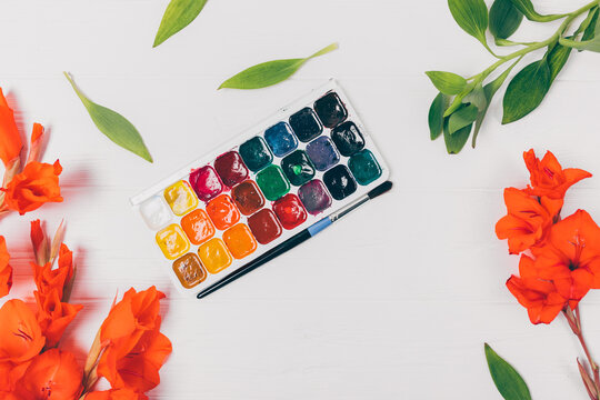 A palette of watercolors is on a table next to some flowers