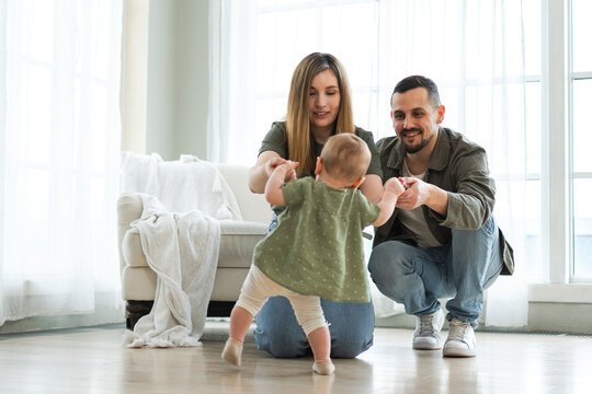 Baby development. Little infant girl making first steps at home. Happy mother father looking at toddler child walking in living room. Happy family at home. Mom dad supporting baby first steps