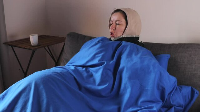 woman wrapped in blue blanket with hood shivers from the cold in her apartment. start of the heating season marks arrival of the cold season. She appears to be unwell.