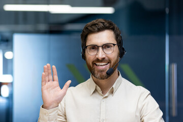 Close-up portrait of a young man in a headset and glasses sitting in the office in front of the camera, greeting and waving