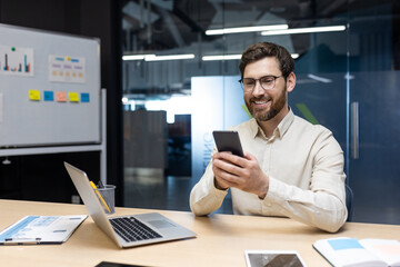 A smiling young man works in the office on a notebook, sits behind a desk on a whiteboard with...