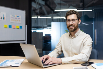 Portrait of a smiling young man sitting in the office behind a laptop and looking smugly at the camera