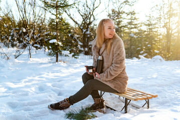 Blonde girl in a fur coat sits on a bench in the winter forest and drinks coffee.