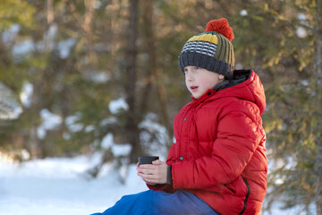 Boy in red jacket and hat sitting on the snow with a cup in his hand, sits and looks into the...