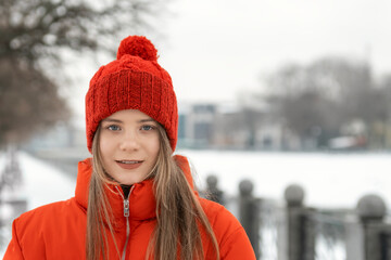 Close-up portrait of blue-eyed long-haired girl in red knitted hat and red jacket on winter nature...