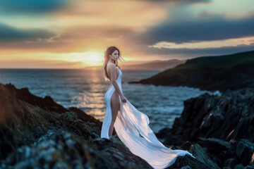 Woman in flowing white dress gazing at sunset on rocky coastal landscape.