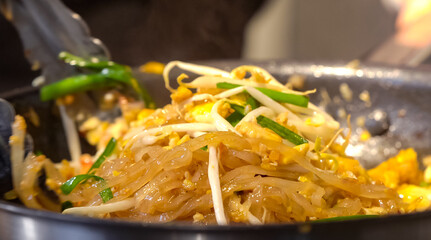 A cook stirs fry Pad Thai noodles in a pan with spatulas on electric stoves. Sauce dishes for cooking traditional Pad Thai.