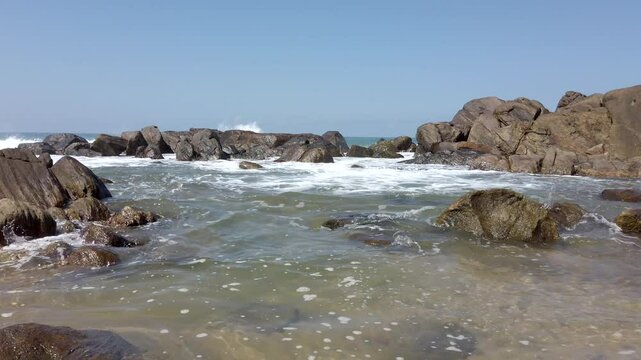 Ocean waves crash over rocks, filling a small tidepool under a bright sun, creating a serene coastal scene