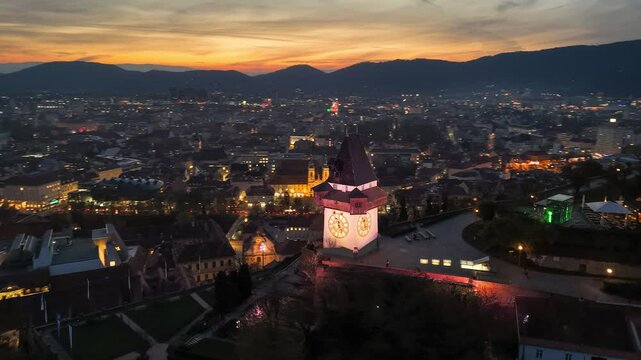 Aerial view of Uhrturm clock tower on Graz's Schloberg hill in the evening. Graz, Austria at dusk with the clock tower illuminated. Vibrant sunset sky at background