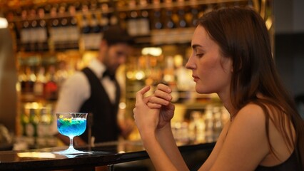 Sitting beautiful woman guest feeling bored waiting friends or couple with drinking mixed vodka in blue hawaii cocktail at luxurious counter bar for Friday meeting on bartender background. Vinosity.