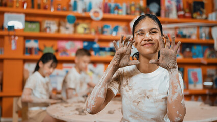 Happy caucasian girl pose at camera while diverse children modeling clay behind. Cute student wearing dirty shirt while looking at camera at workshop in art lesson. Blurring background. Edification.