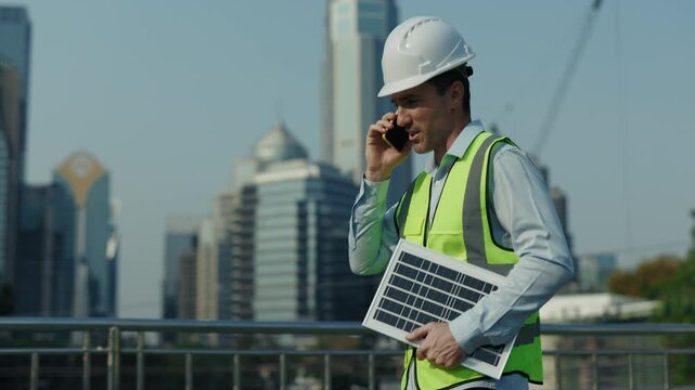 Portrait of busy man engineer talking on smartphone walking along footbridge against high-rise office buildings. Male worker holds solar panel offering innovative technology to client