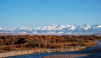 A serene river meanders through a rugged landscape, flanked by autumnal shrubbery, leading the eye to the majestic snow-covered mountains under a deep blue sky