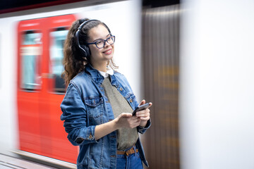 Pretty young girl listening to music with headphones at the train station while holding her phone