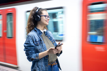 Pretty young girl listening to music with headphones at the train station while holding her phone