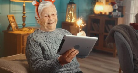 Smiling cheerful old grandmother with sparkler stick at home celebrating Christmas or New Year having video call on digital tablet with family - Powered by Adobe