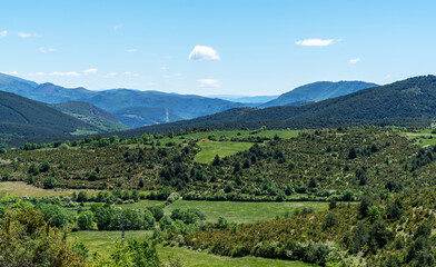 A panoramic view stretches over a verdant valley dotted with trees, sweeping towards rolling hills and distant mountains under the vast expanse of a clear blue sky.