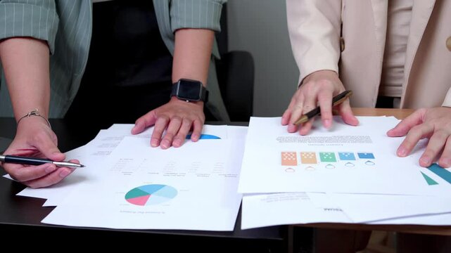 Close-up of two people's hands reviewing financial documents. A pie chart displays "% Income Per Product," and a table lists costs, profits, and returns. One hand holds a pen