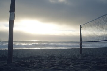 A volleyball net stands on a sandy beach with the ocean and sunset in the background.